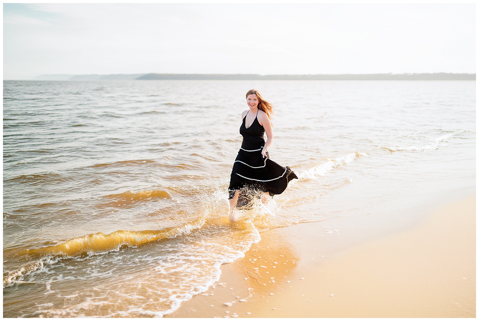 High school senior splashing through ocean water in black maxi dress from Star City Boutique at beach.